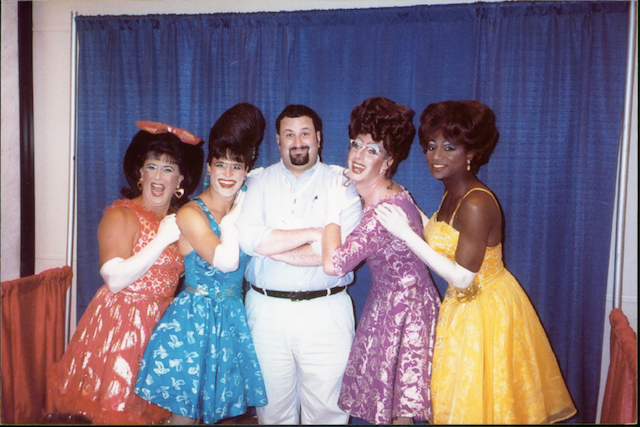 david-and-kinsey-sicks David poses with the four members of the Kinsey Sicks, dressed in colorful dresses, elaborate wigs, and white gloves, in front of a blue curtain.
